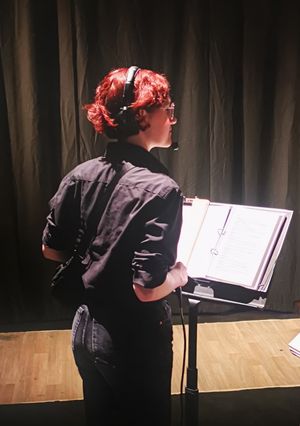 Louisa is wearing a headset with her back to the camera. In front of her on a music stand is a script.